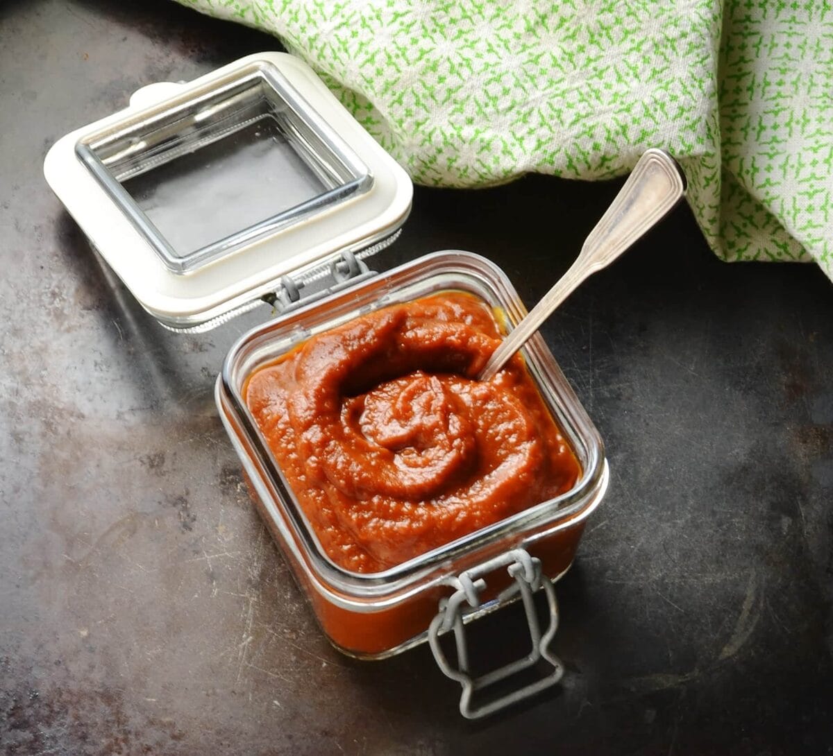 Barbecue sauce in square jar with spoon and green cloth at the back on, on dark brown surface.