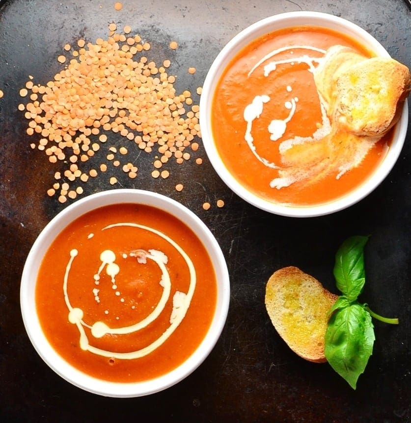 Top down view of tomato soup in 2 white bowls with toasted bread, lentils and basil leaves on oven tray.