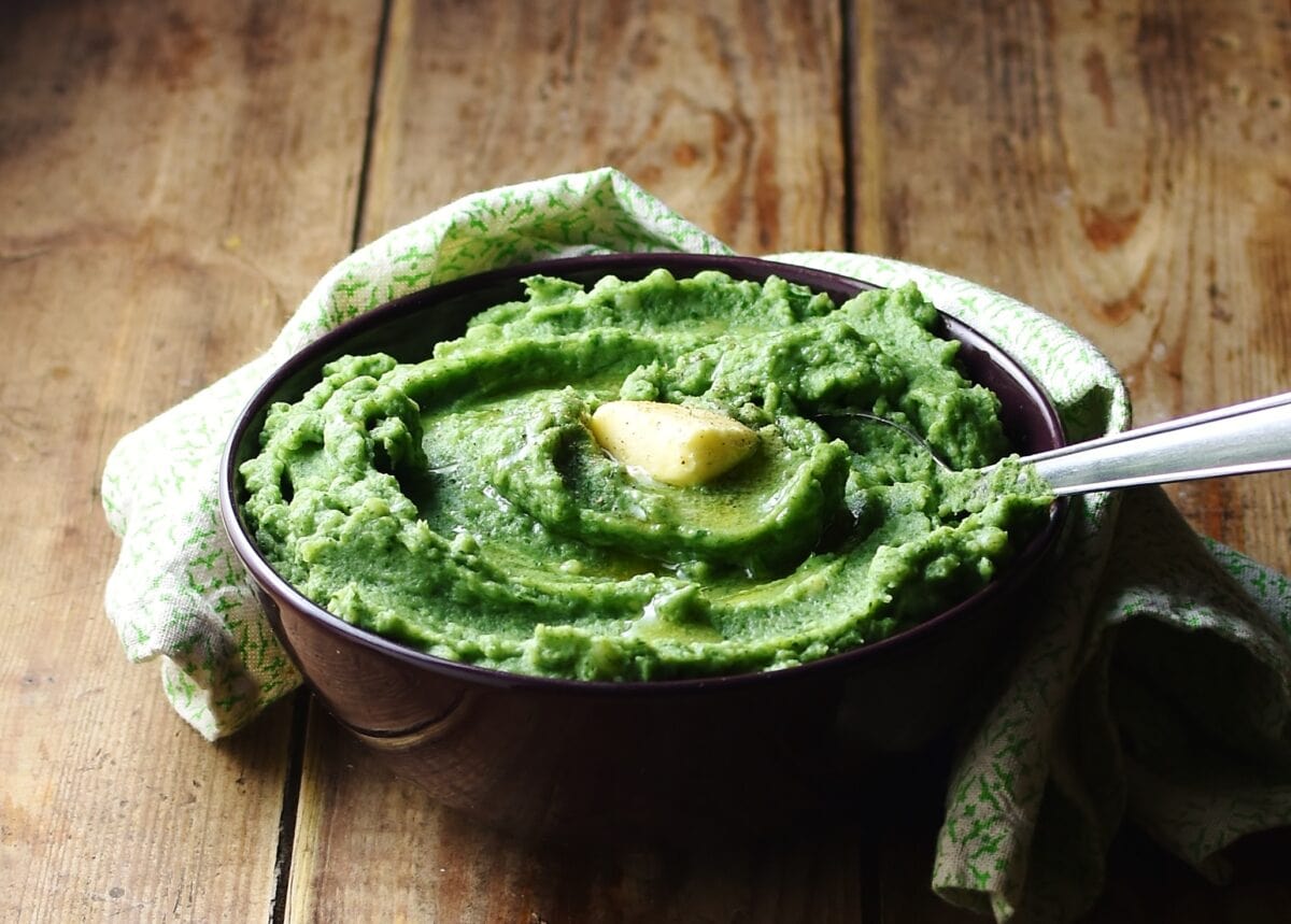 Side view of spinach mashed potatoes with knob of butter and spoon in purple bowl wrapped in green cloth.