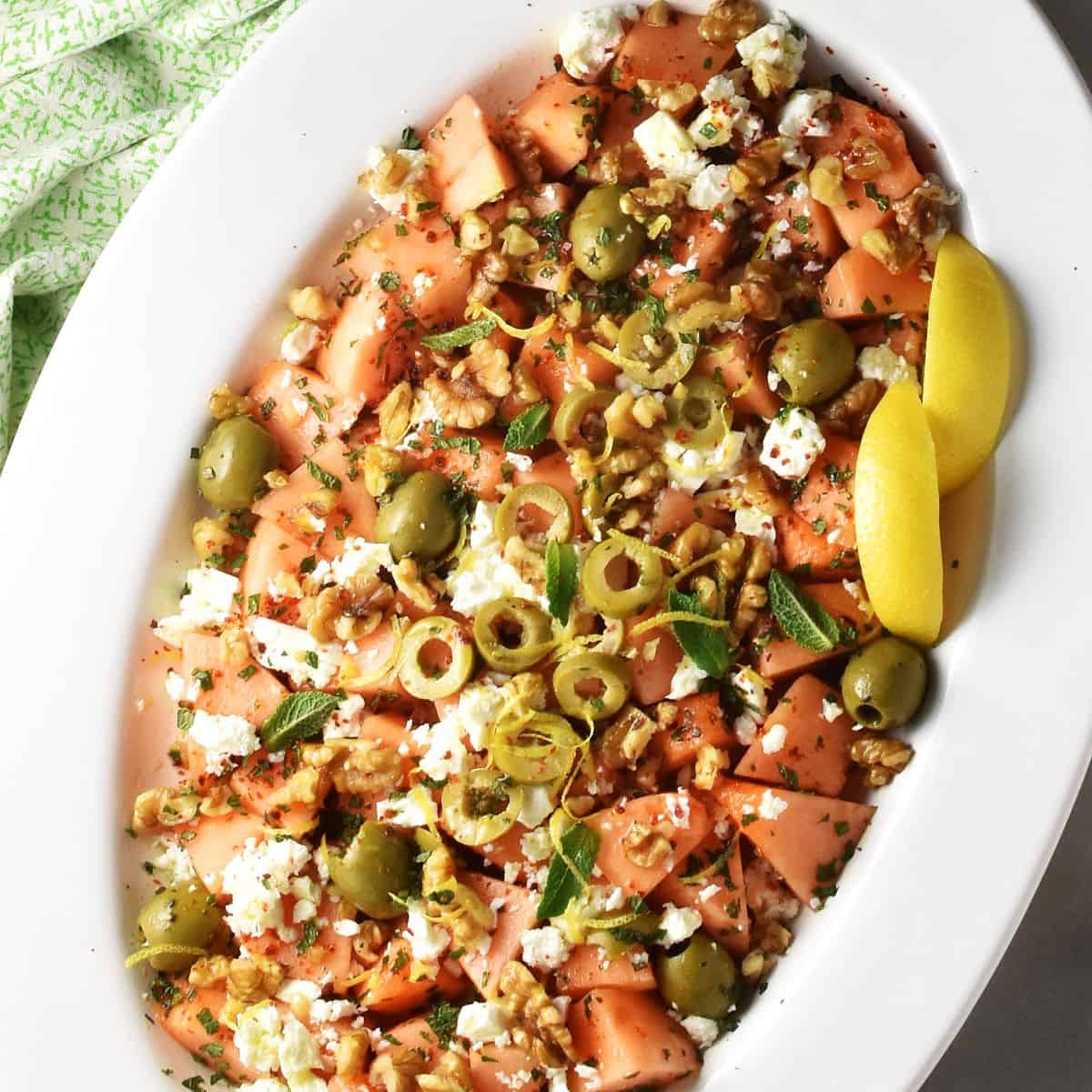 Top down view of cantaloupe salad with feta, olives and lemon wedges on white oval platter.