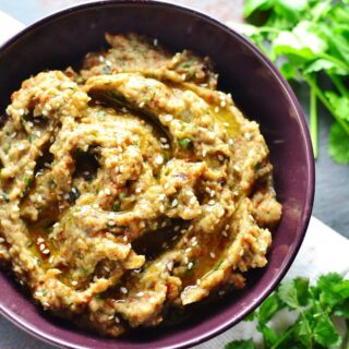Eggplant dip in purple bowl on white cloth with fresh cilantro in top and bottom left corners.