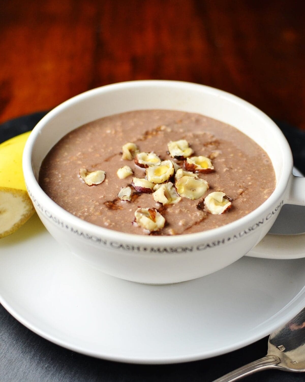 Mocha overnight oats topped with crushed hazelnuts in coffee cup on top of saucer with half banana in background.