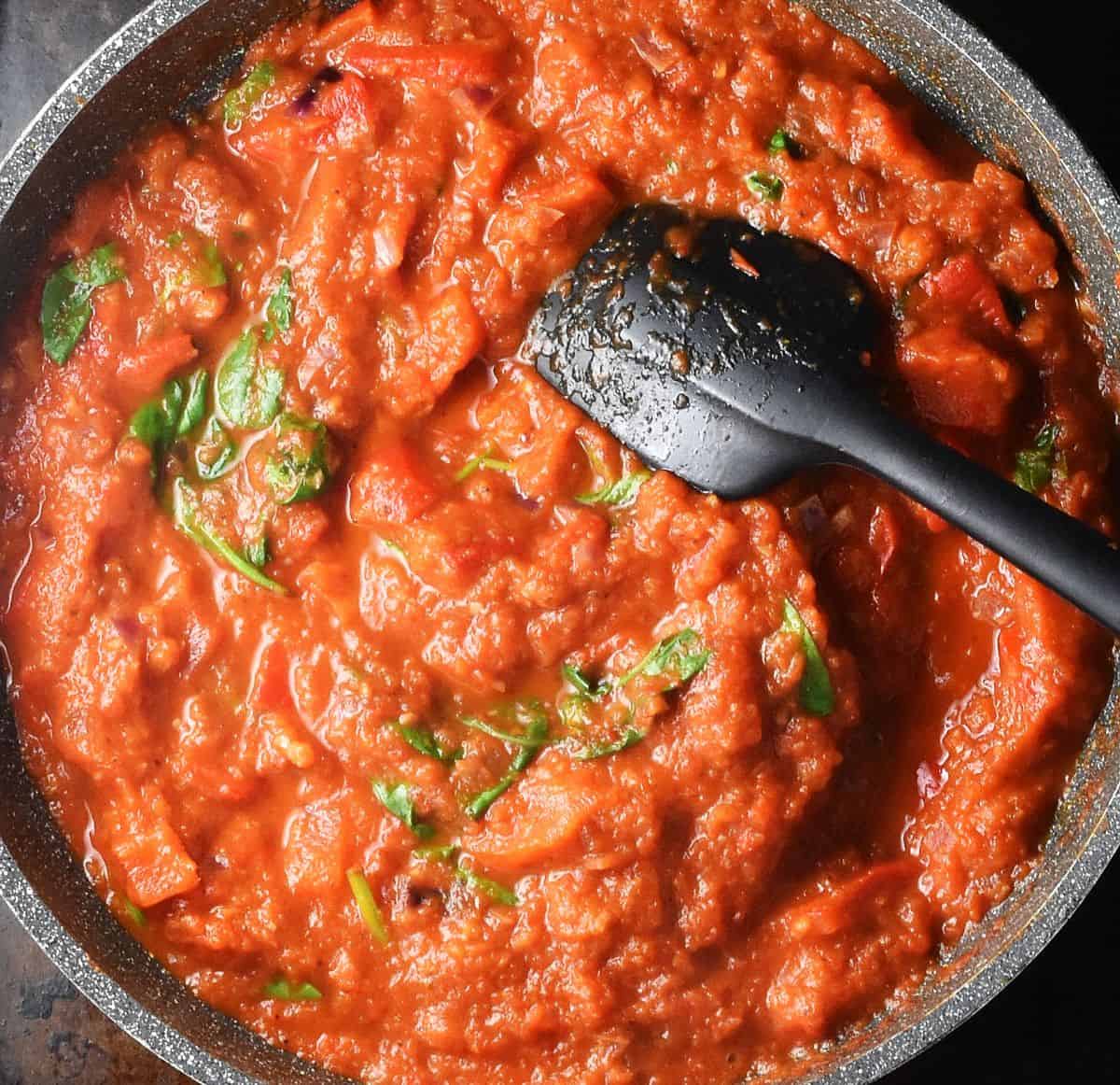 Thick sauce with tomatoes, peppers and spinach in large pan with black spoon.