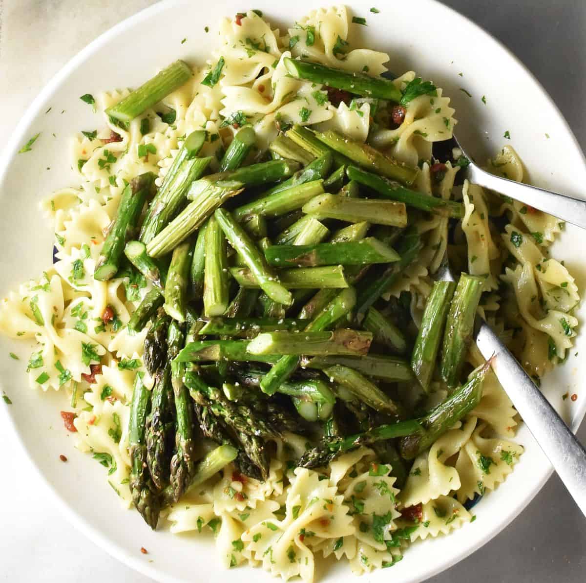 Pasta with chopped roasted asparagus tips in white bowl with 2 spoons.