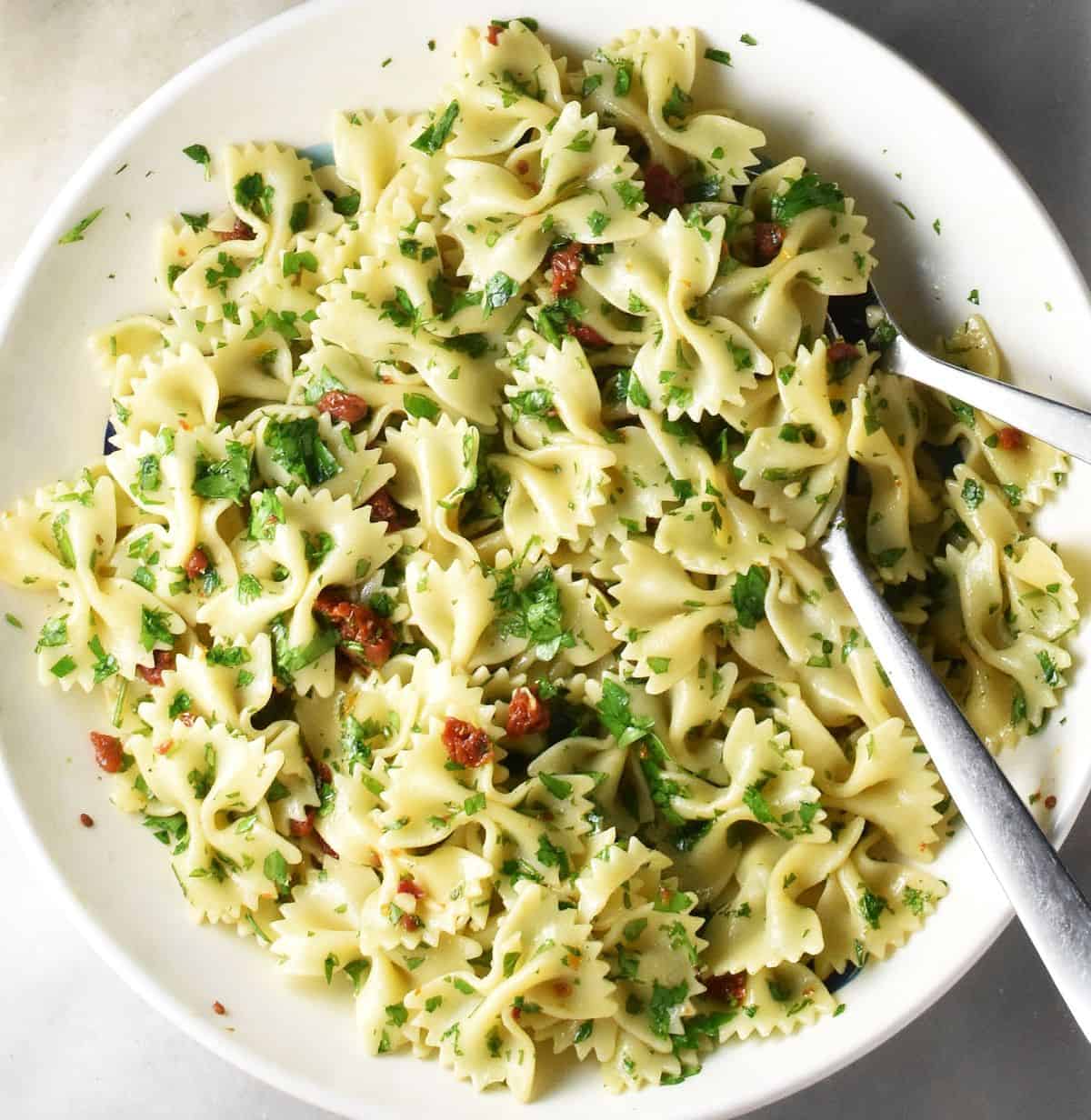 Top down view of farfalle pasta with herbs and 2 spoons in white bowl.