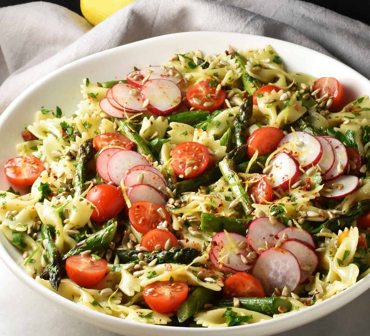 Side view of pasta salad with asparagus, tomatoes and radishes in shallow white bowl.