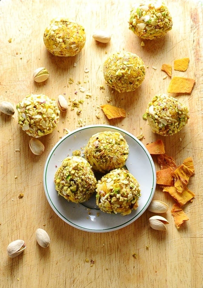 Mango cheesecake bites on small white plate and wooden board with dried mango pieces and pistachios scattered about.
