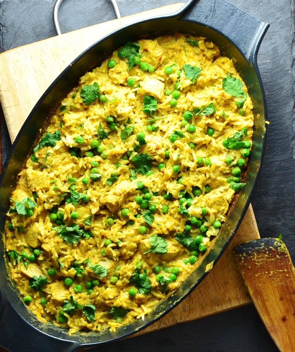 Top down view of curried chicken rice in black oval cast iron dish on top of wooden board with wooden spoon in bottom right corner,