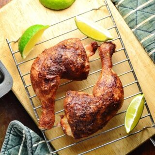 Top down view of 2 crispy chicken leg quarters on top of wire rack with lime wedges on top of wooden board.