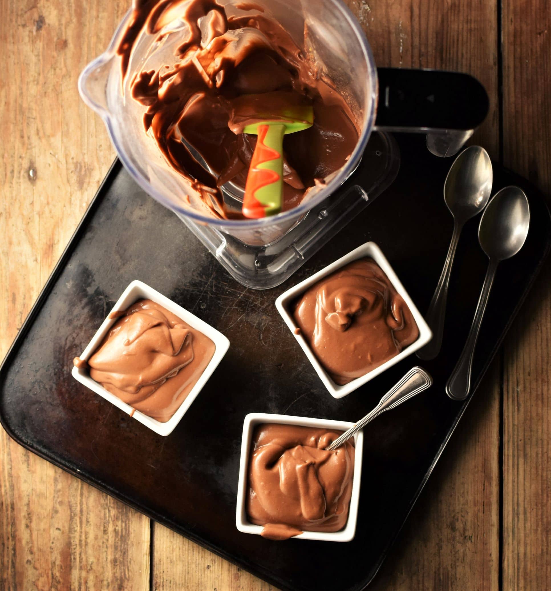 Chocolate pudding in 3 white square dishes and in blender with spatula, plus 3 spoons on top of black tray.
