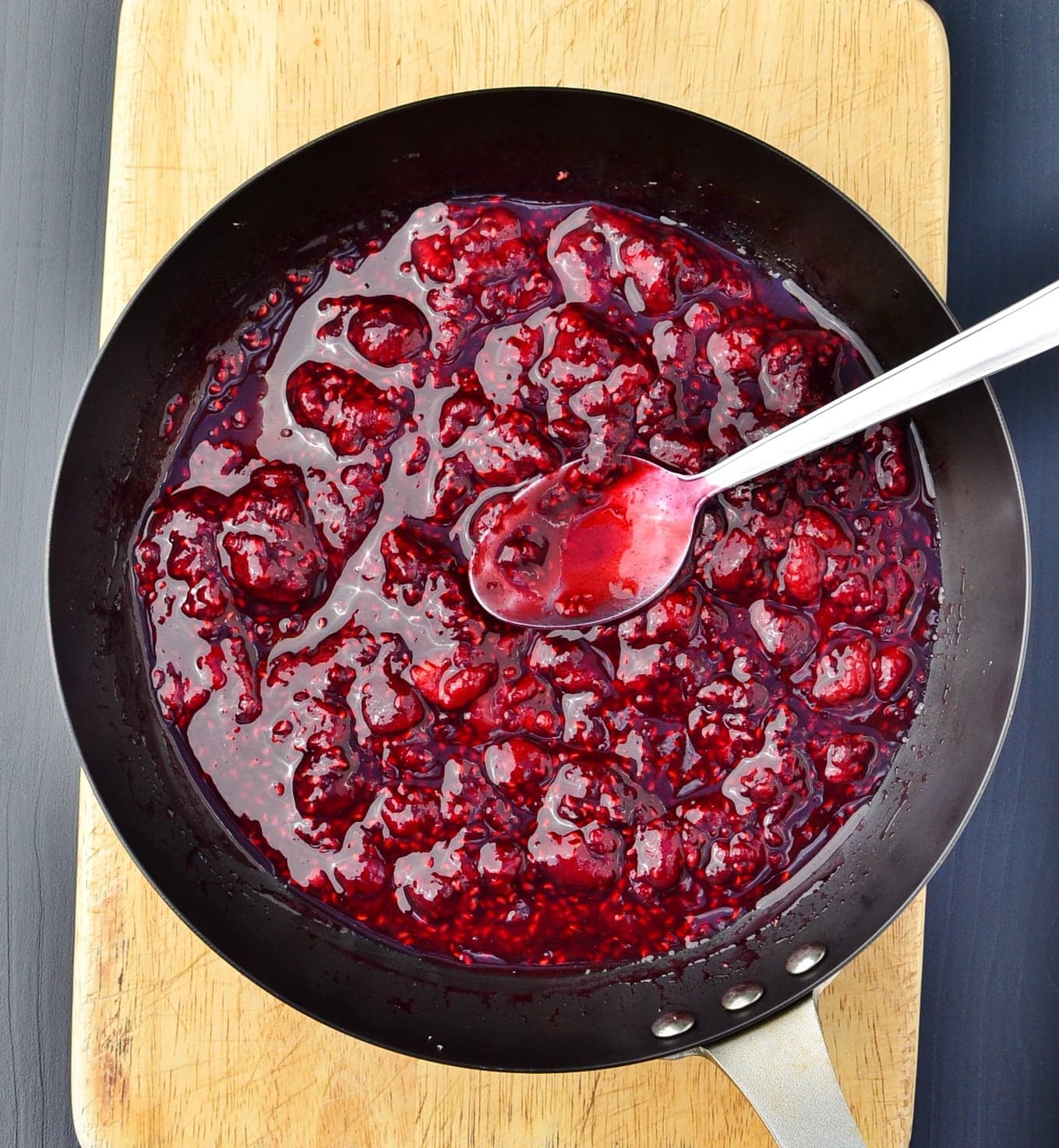 Top down view of cooked raspberries with spoon inside skillet on top of wooden board.
