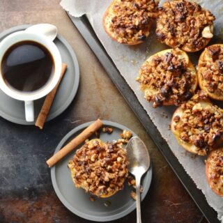 Top down view of apple halves with browned pecans on top of baking sheet and grey saucer, with coffee in white cup and grey saucer.