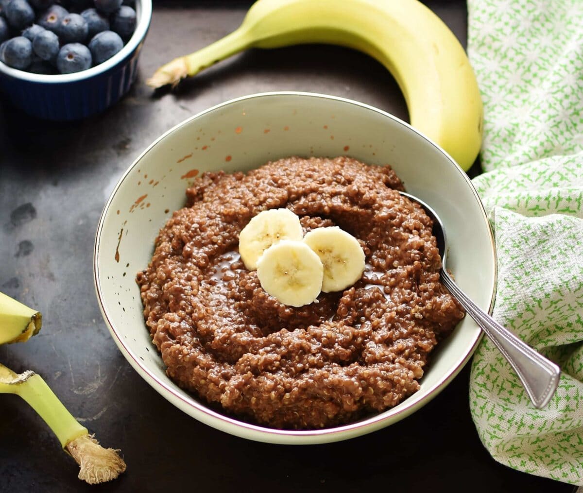 Quinoa chocolate porridge with slices of banana and spoon in light green bowl, with green cloth, banana and blueberries in small dish in background.