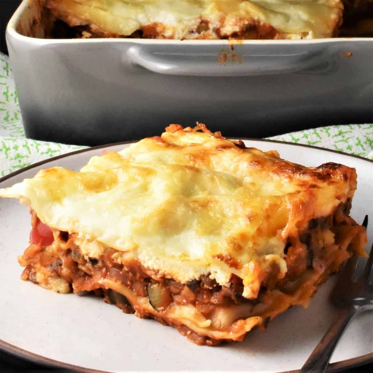 Side view of lentil lasagna piece on white plate with grey casserole dish in background.