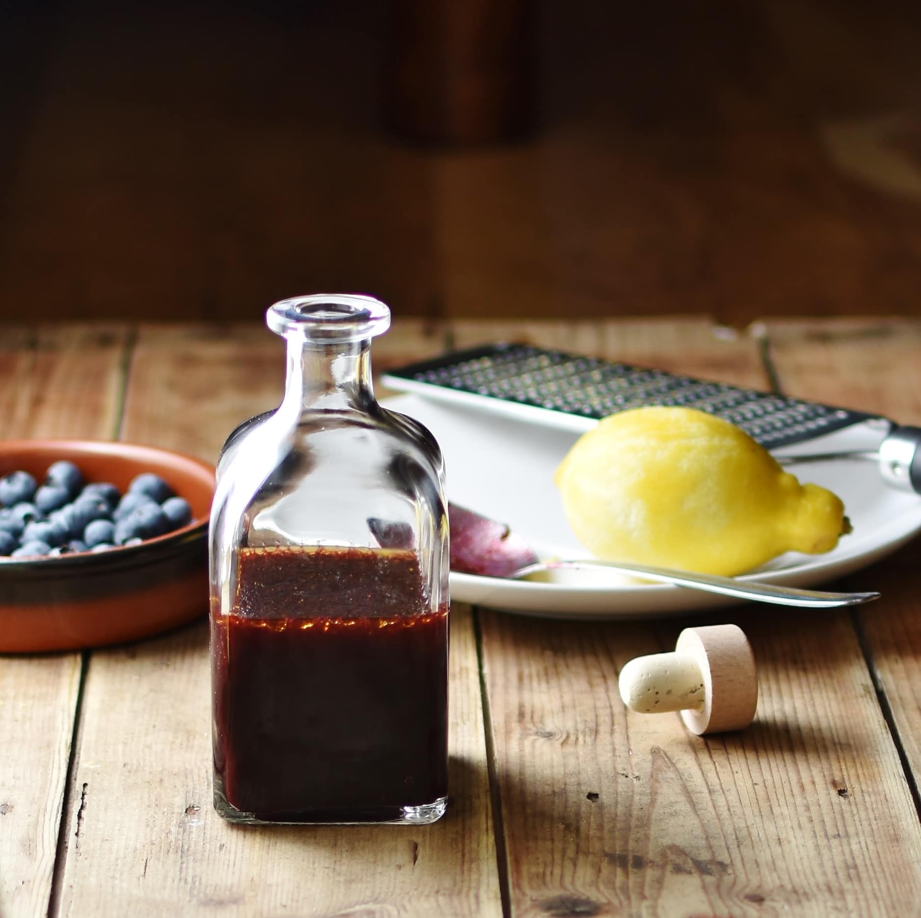Blueberry dressing in bottle, with blueberries in brown dish, lemon, zester and spoon on top of white plate and bottle top in background.