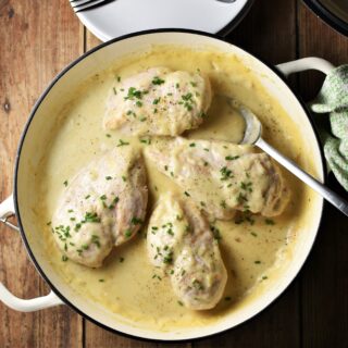 Top down view of 4 chicken breasts in white sauce with spoon in large white pan, with green cloth to the right and plates at the top.