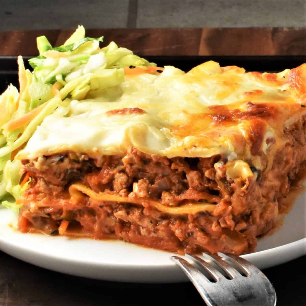Side view of turkey lasagna slice with slaw and fork on white plate.