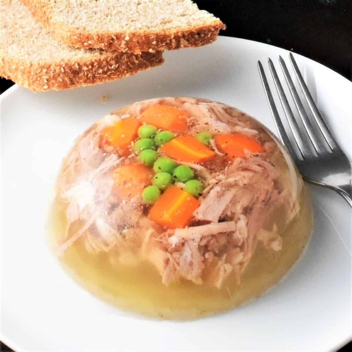 Side view of meat jelly on white plate with fork and bread in background.