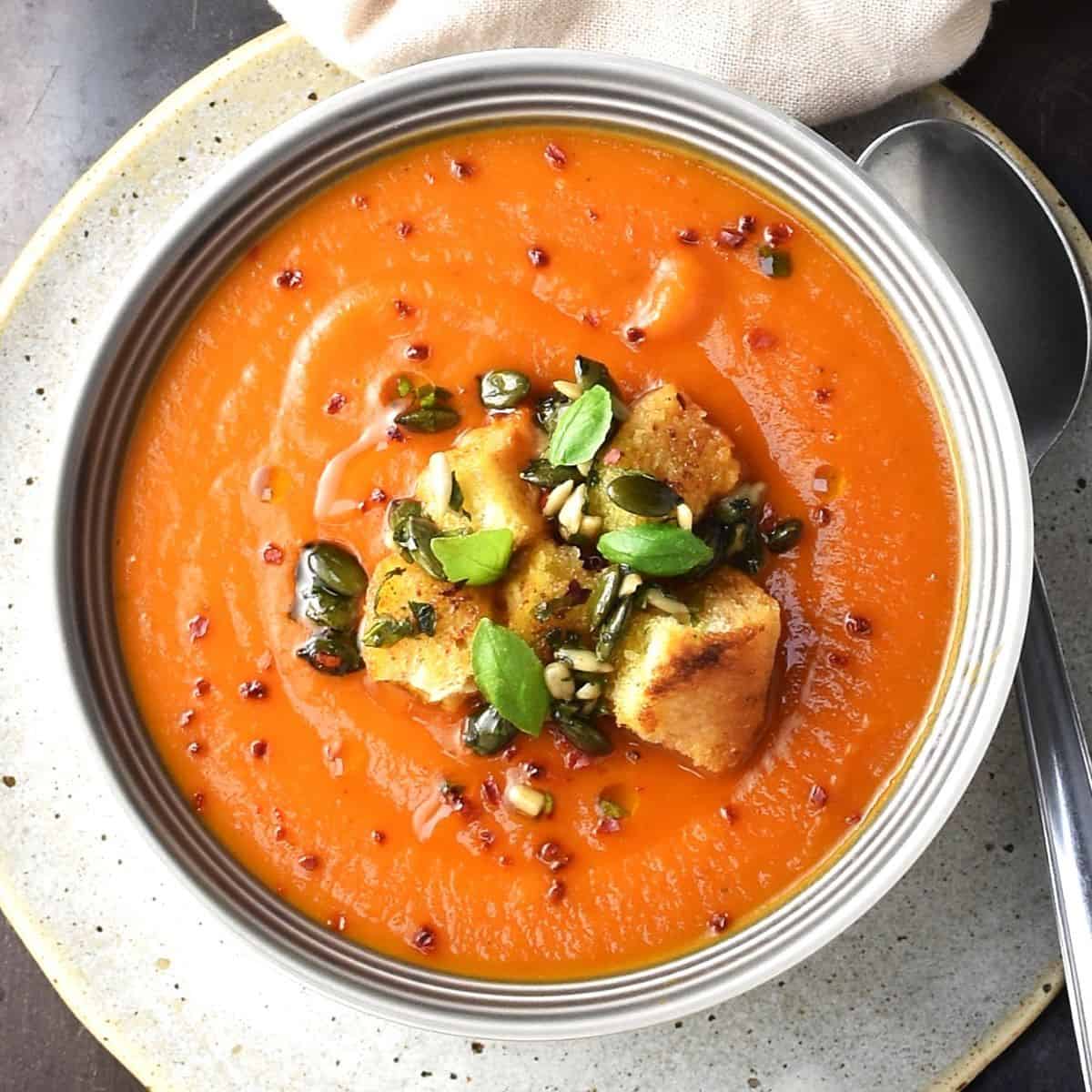 Top down view of creamy butternut squash tomato soup with croutons in grey bowl with spoon on right.
